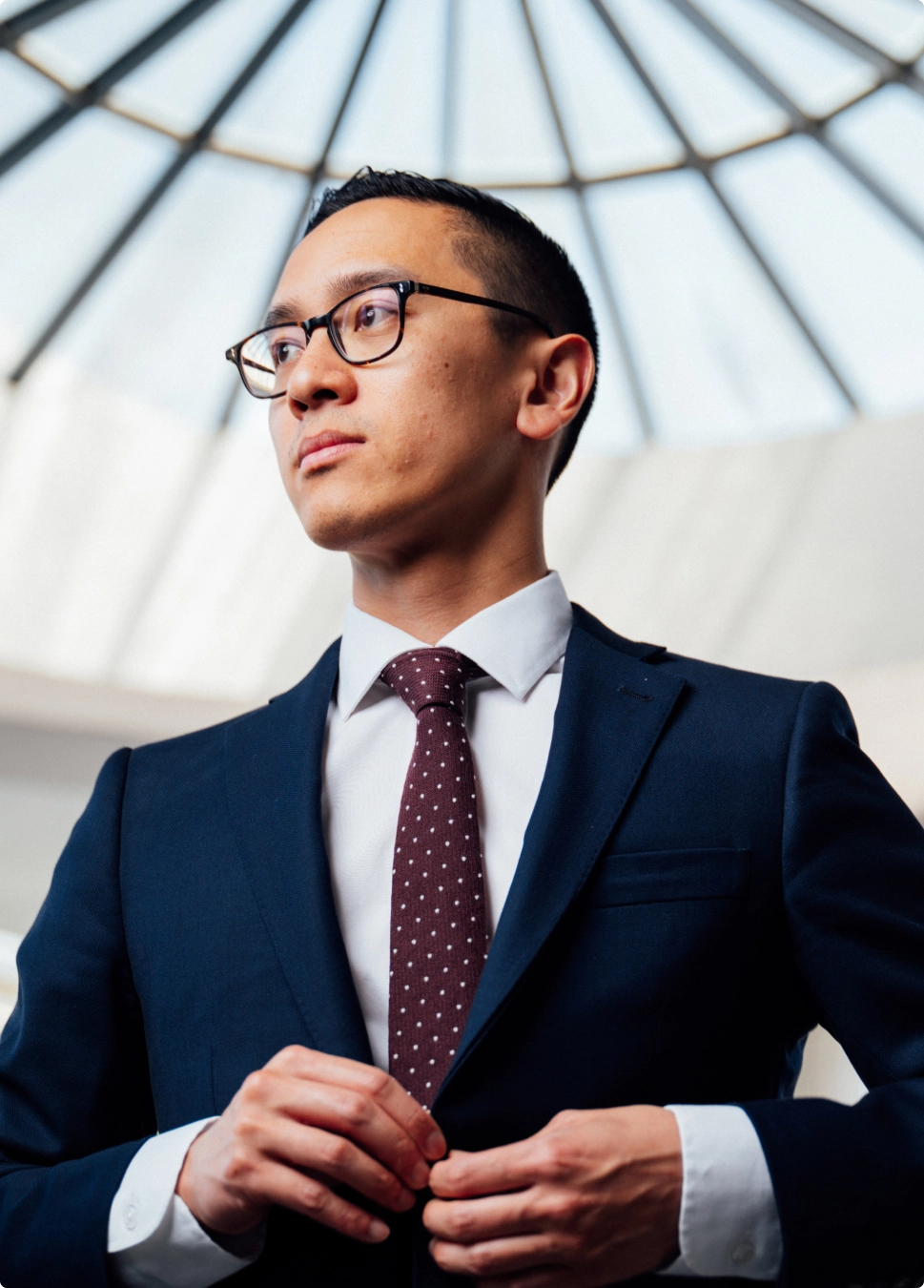Professional businessman in navy suit and burgundy tie adjusting jacket under modern glass dome architecture