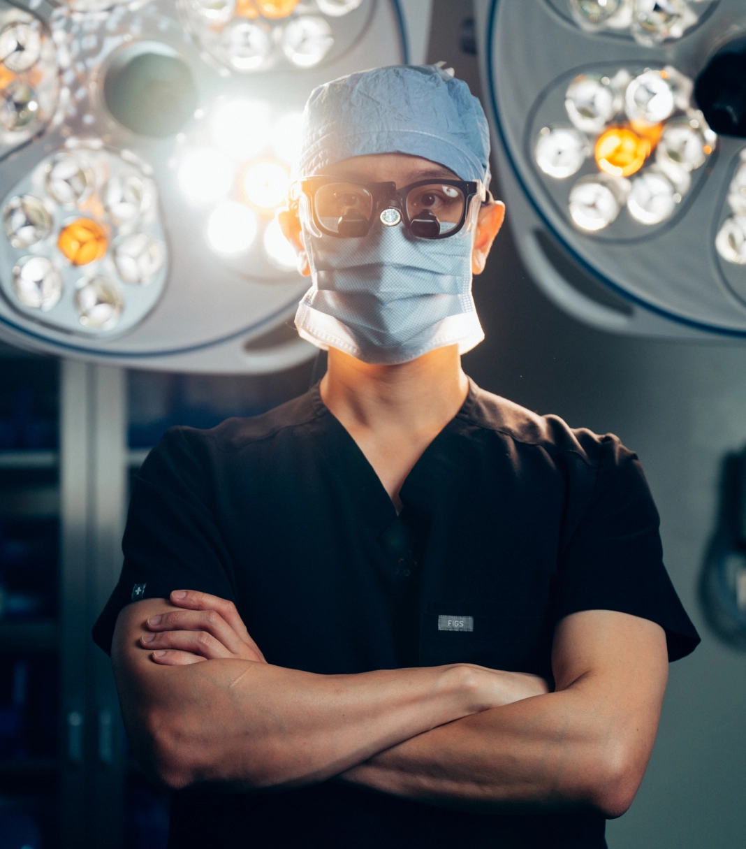 Surgeon in scrubs and mask standing with crossed arms in operating room with surgical lights overhead