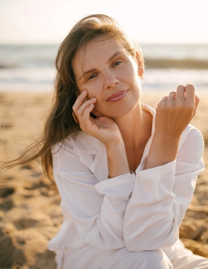 Woman in white shirt smiling on beach at sunset with hand near face in natural light portrait