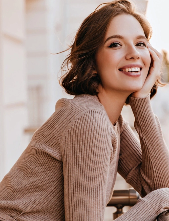 Smiling woman with auburn hair wearing beige ribbed sweater resting chin on hand in soft natural light