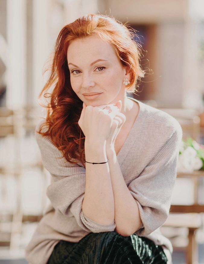 Red-haired woman in beige sweater with hands clasped near face, soft natural lighting portrait