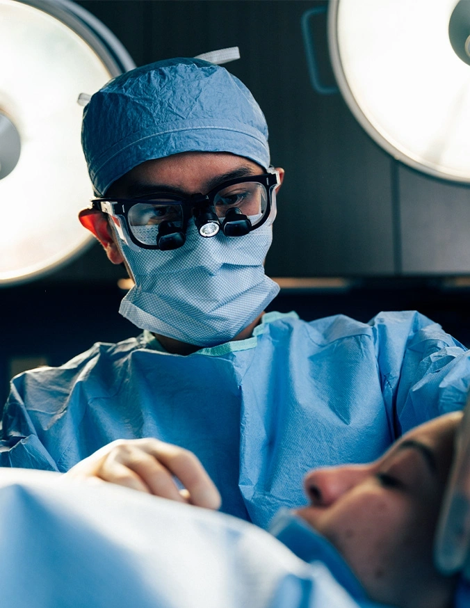 Surgeon in blue scrubs and mask performing surgery in operating room with surgical lights overhead