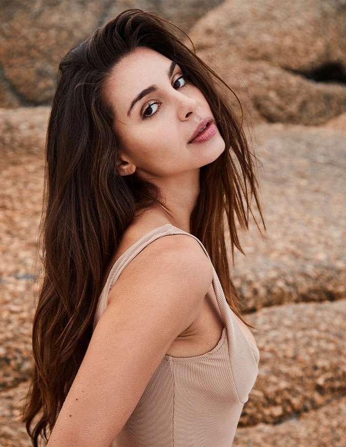 Woman with long dark hair in beige tank top posing outdoors against natural rock formation background